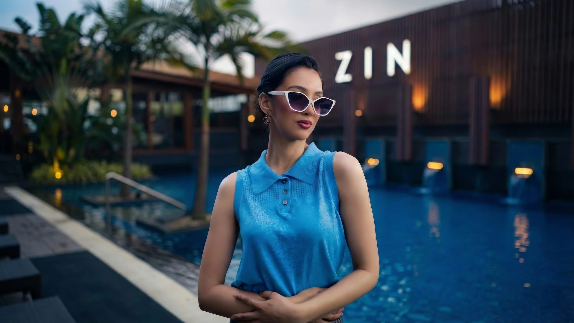 Woman in blue polo and sunglasses posing by a luxury pool at dusk.