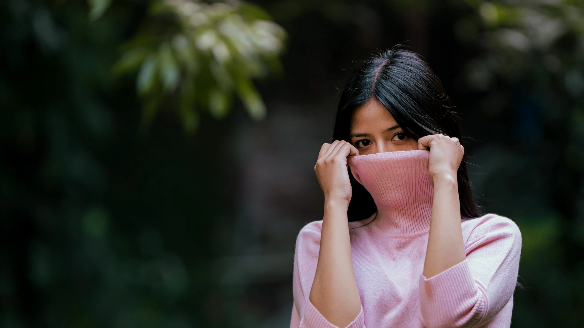 Woman in pink turtleneck partially covering her face against dark foliage.